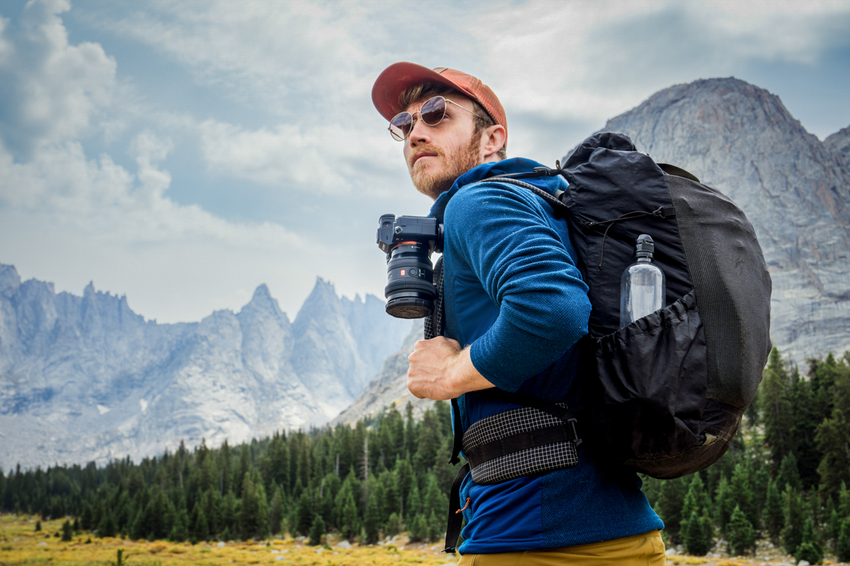 A man hiking with a camera on his backpack strap in front of mountains