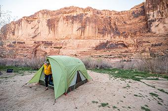 A camper pitches a spacious tent against a desert background of sheer, red rock walls. 