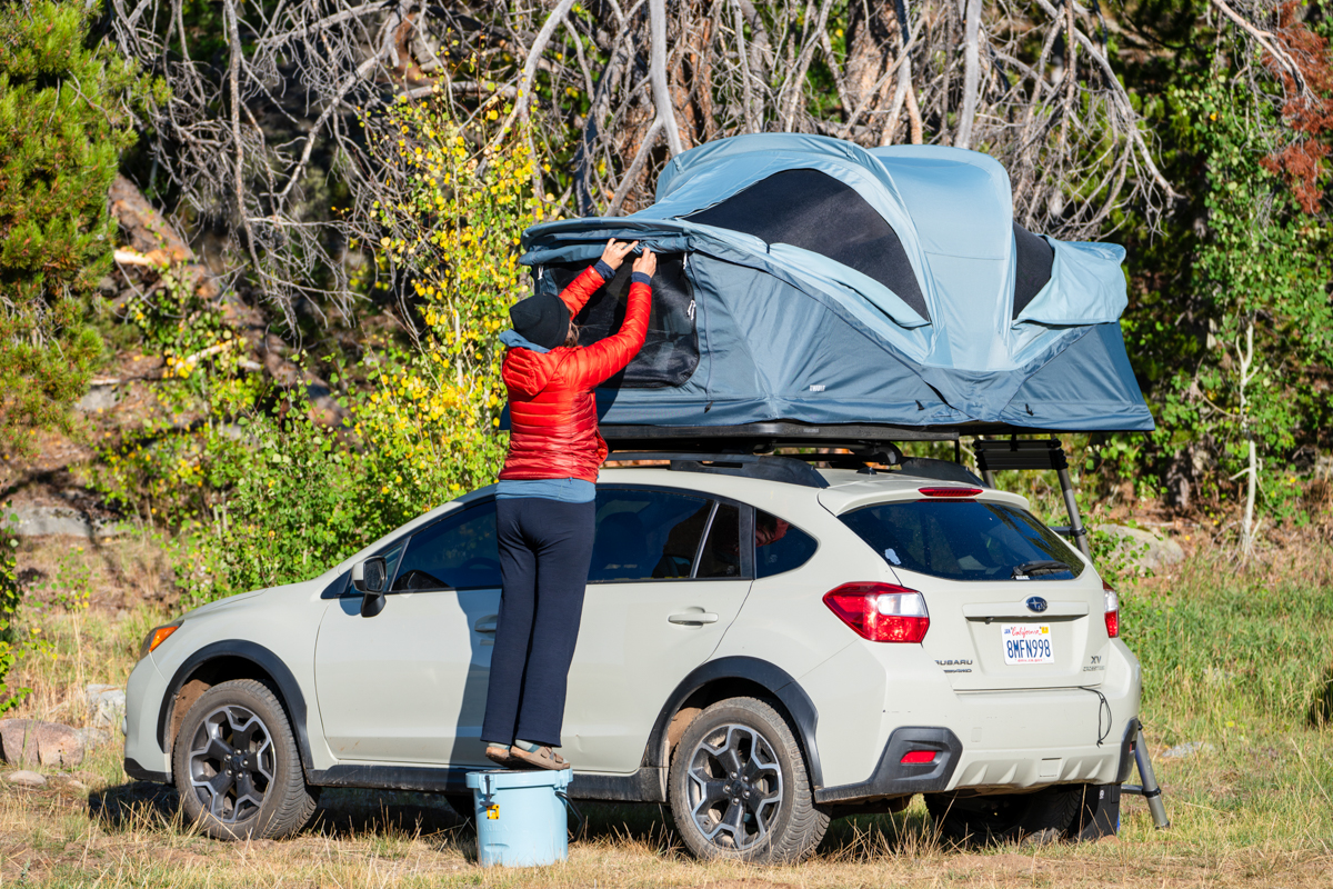 A woman standing on a cooler to adjust a rooftop tent