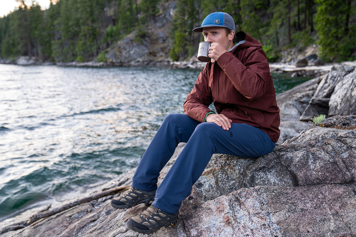 A hiker has a cup of coffee while sitting on a rock.
