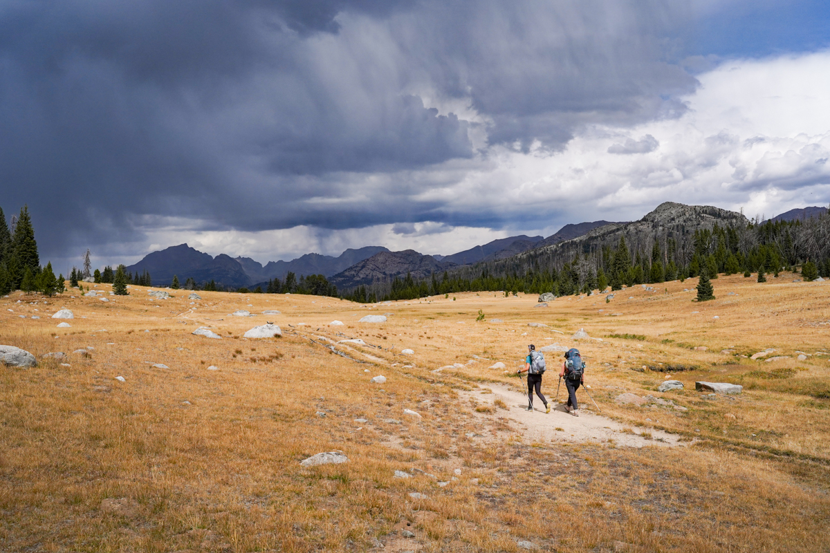 Two hikers cross a long, empty field in the mountains.