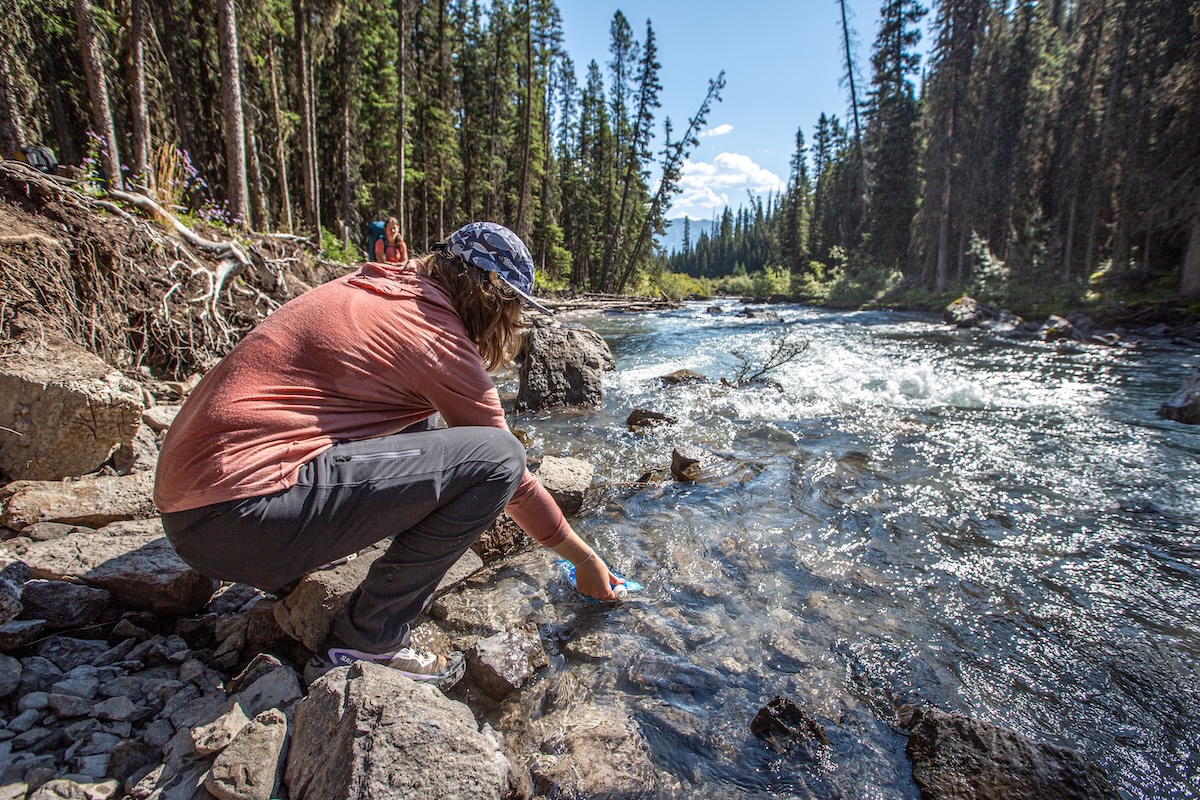 A hiker stops to fill a water bottle in a stream.
