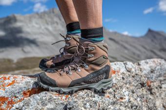 A closeup of a pair of hiking boots standing on a rock with mountains in the background