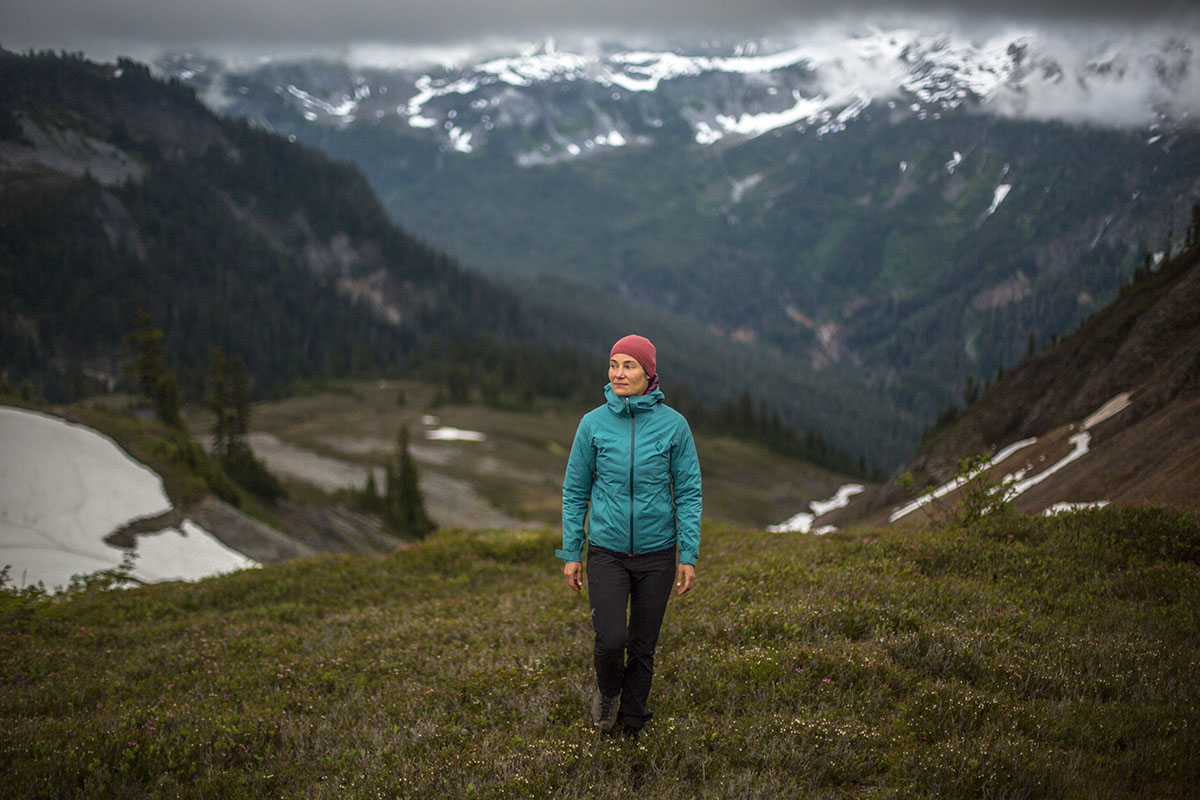 A woman hikes the valley between two mountains, wearing her hat and a rain shell.