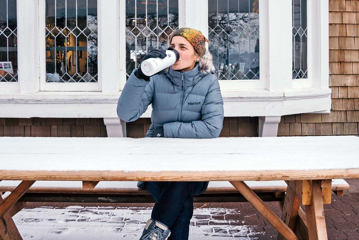 A woman drinks hot chocolate from a thermos outside on a winter day.