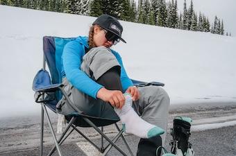 A woman sits in a camping chair in a ski resort parking lot pulling on her socks