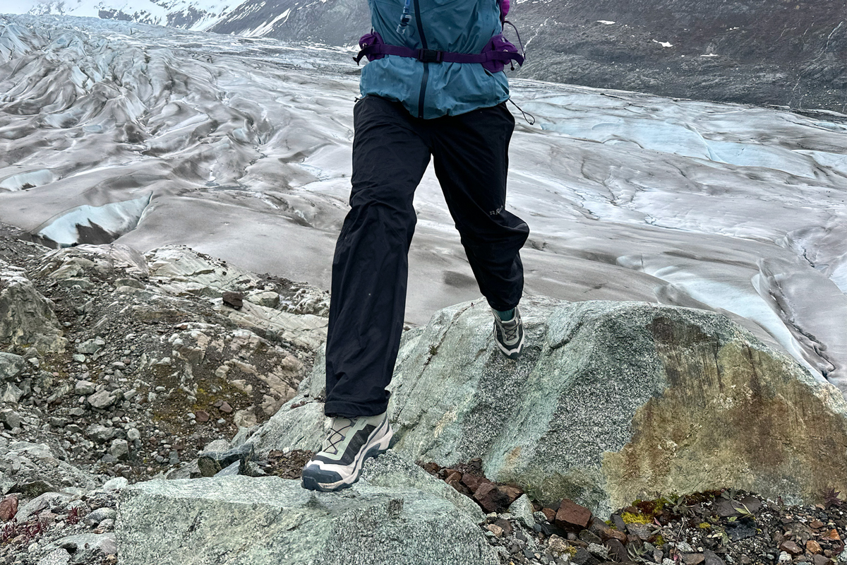 A hiker stepping down onto a boulder in hiking shoes while hiking in a rocky and glaciated landscape