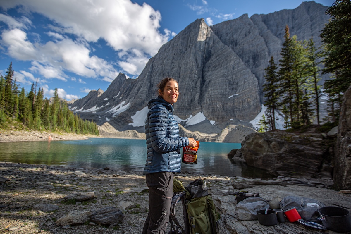 A hiker unpacks her backpack next to a high granite peak and blue alpine lake.