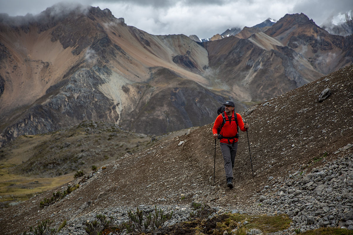 A hiker treks up a distant, high-mountain trail with soaring, sandy peaks in the background.