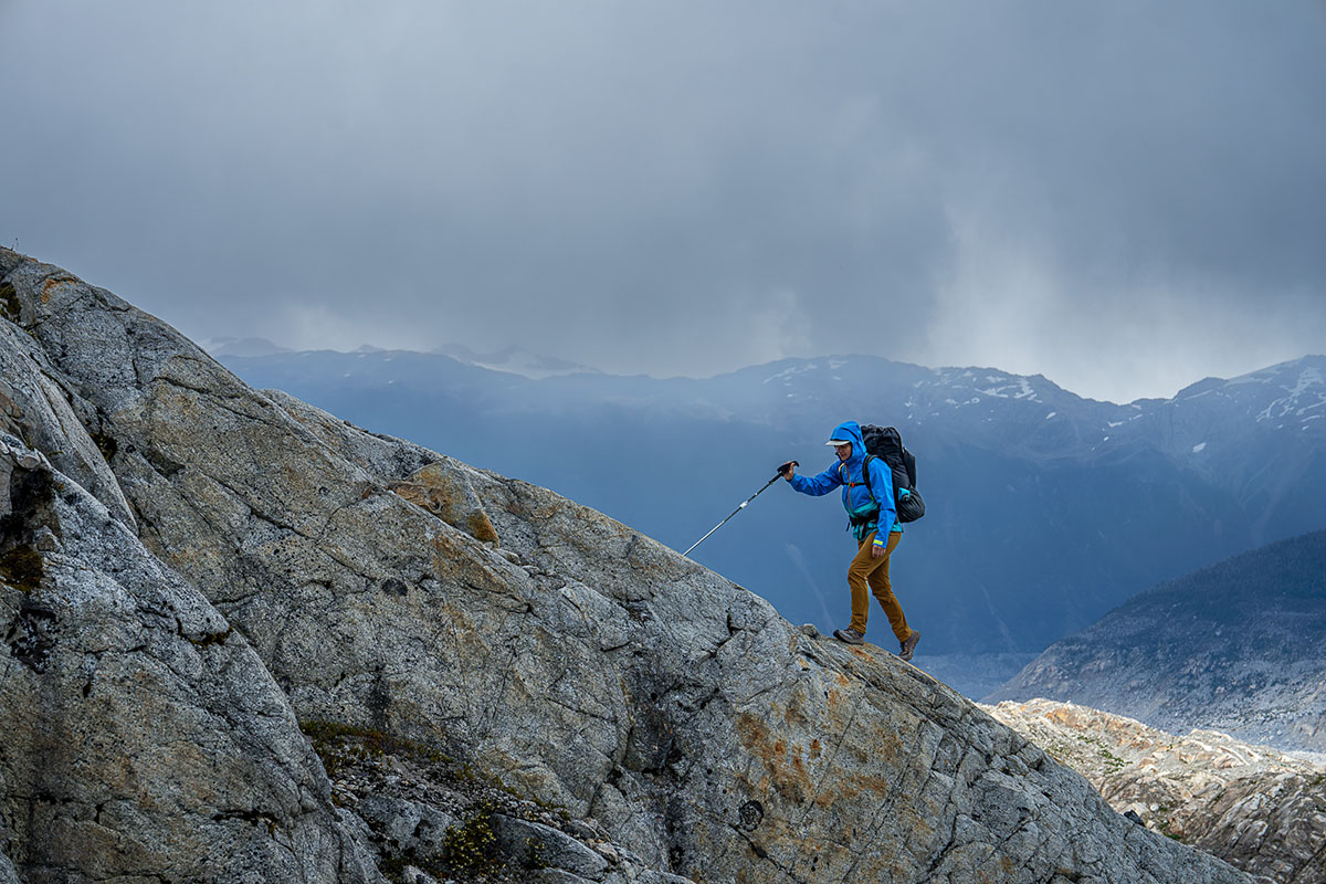 A hiker extends their hiking pole in front of them to navigate a rocky granite ridge in the mountains.