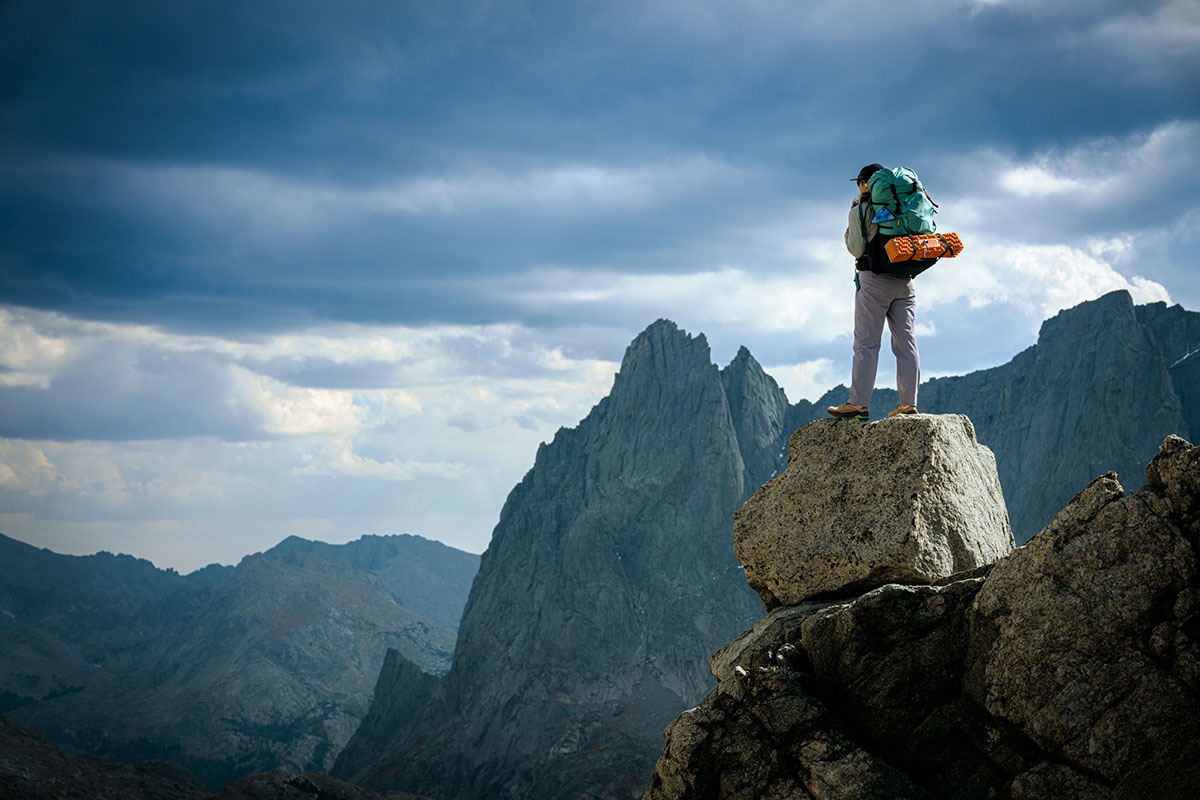 A woman stands on a rock with her backpacking backpack, taking in an epic mountain view.